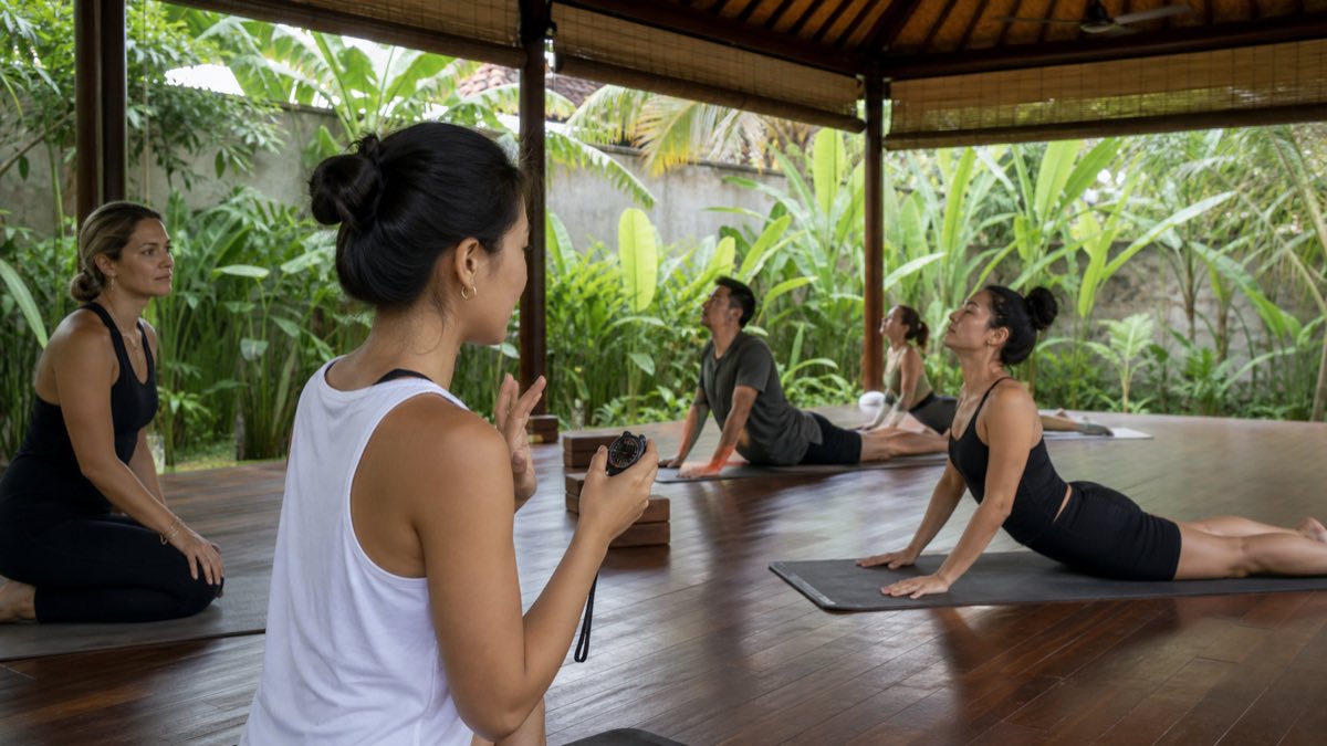 International trainees practising verbal cues and sequence timing with a stopwatch inside a naturally heated tropical yoga studio in Bali