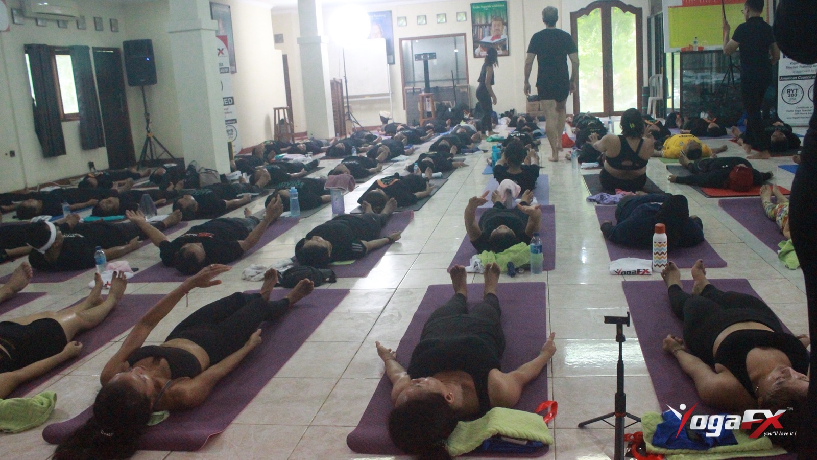 Students in a hot yoga class lying on purple mats in final relaxation while the teacher walks the room